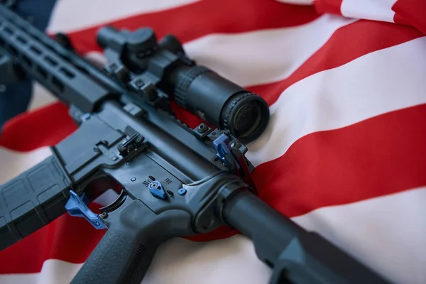 Modern gun equipped with optical scope resting on flag of the USA