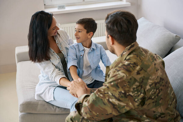 Woman and kid smiling at each other while sitting in front of army man on sofa and holding his hand