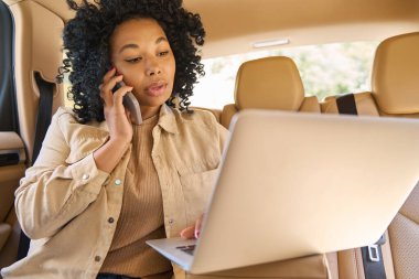Young mixed race woman is absorbed in urgent work at a laptop and phone in the back seat of car