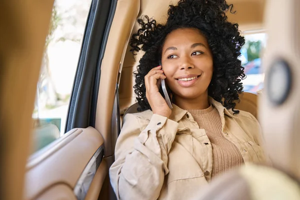 Curly young female travels comfortably in the back seat of a car and talks on the phone with a smile