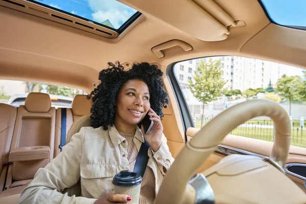 Joyful woman with morning coffee in her hands communicates cutely on the phone in the interior of a comfortable car