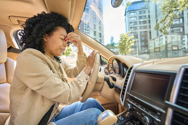 Multiracial woman suffering from pain pinches bridge of her nose, leaning on steering wheel in interior of a beautiful car