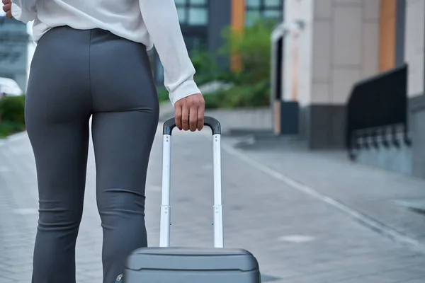 Woman in gray trousers walks down the street with a small suitcase on wheels