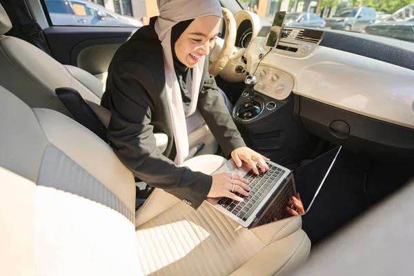 Woman in a hijab and a headdress works on a laptop in a car
