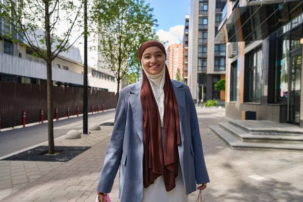 Woman in a hijab walks along a sunny street with purchases in her hands
