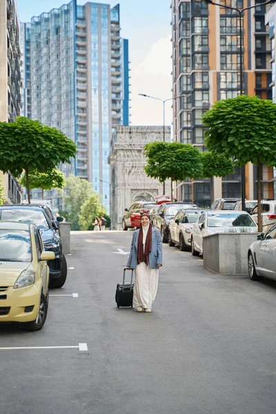Arab woman in a hijab walks along the street against the backdrop of high-rise buildings with a suitcase on wheels