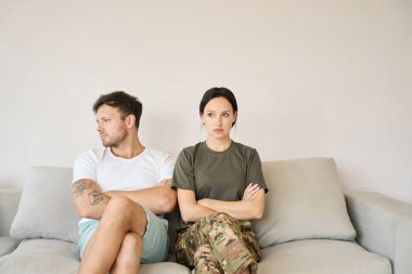 Conflicting couple is sitting on a couch in closed poses, woman in camouflage clothes, man looking dejectedly to the side