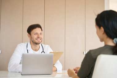 Optimistic doctor communicates kindly with a young patient in a bright office, tablet and a laptop are on the table