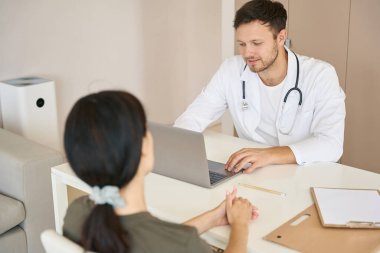 Attentive, kind doctor with a phonendoscope, at his desk, interviews patient about health problems and writes down information in laptop