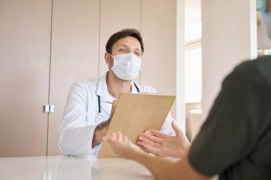 Doctor in a white coat and a protective mask listens attentively and records the patient complaints in writing
