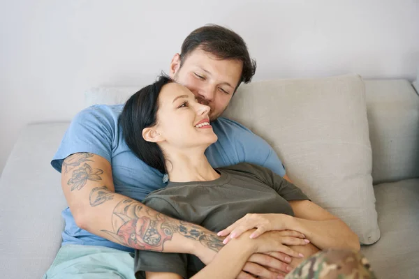 Smiling couple resting on sofa, man gently hugs his girlfriend, the woman is joyful in the arms of her boyfriend