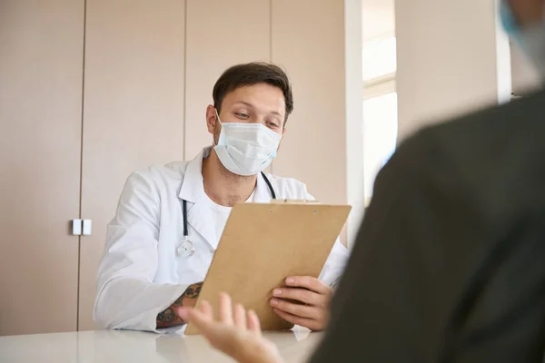 Young doctor in bright room, with a statoscope and in protective mask, consults patient and looks at the medical history