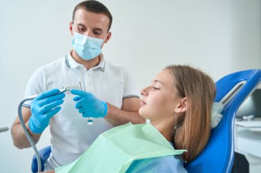 Serious pedodontist holding dental drill handpiece before calm adolescent girl lying in chair