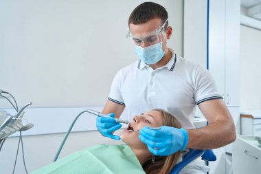 Pediatric dentist using mouth mirror and dental drill during young patient cavity preparation