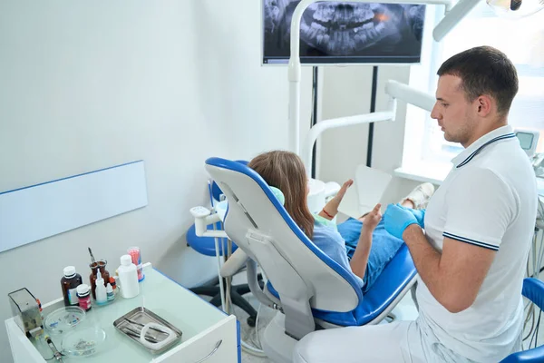 Girl examining her teeth in mirror in presence of serious male stomatologist