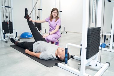 Male patient lying on exercise mat and using medical equipment while doing physiotherapy rehabilitation gymnastics with physiotherapist