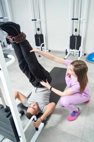 Male patient lifting legs and using medical sports equipment while doing physiotherapy gymnastics with woman physiotherapist