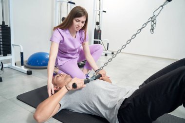 Male patient using medical sports equipment while performing kinesiotherapy gymnastics with woman physical therapist