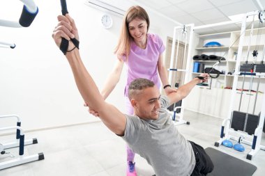 Happy African American man using elastic resistance suspension ropes while performing rehabilitation exercise with physiotherapist assistance