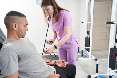 Female medical worker using manual aneroid sphygmomanometer and stethoscope while checking man blood pressure