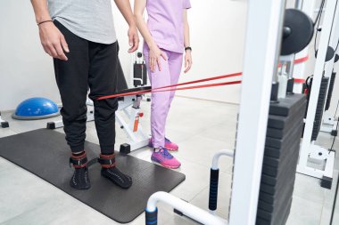 Male patient using kinesiotherapy training equipment while having rehabilitation workout with doctor after leg injury