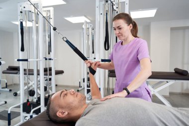 Male patient using kinesiotherapy training equipment while having rehabilitation workout with woman physiotherapist