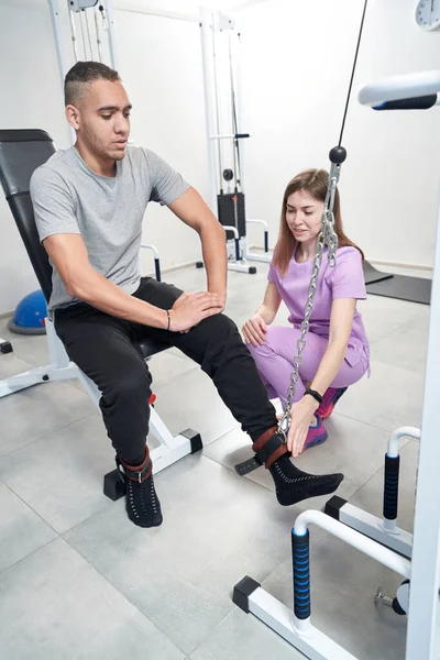Male patient using medical physical therapy equipment while doing rehabilitation exercise with physical therapist