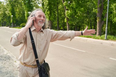 Joyful grey-haired man raising his arm and keeping telephone near ear during conversation