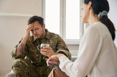 Dejected young military man at the reception of woman psychologist in bright office, the specialist listens attentively to the patient