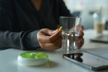 Multiracial female sits at a table and holds a yellow oblong pill and a glass of water in her hands
