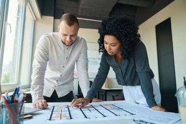 Enthusiastic office staff, multiracial woman and a caucasian man discussing working draft near professional blueprints