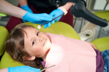 Calm little patient lying in chair in presence of pediatric dentist and assistant during check-up