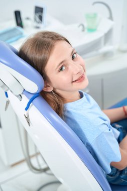 Cheerful young female patient sitting in dental chair and smiling at camera
