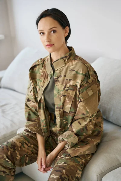 Charming pretty woman military in a camouflage suit sits on a soft sofa in a bright room