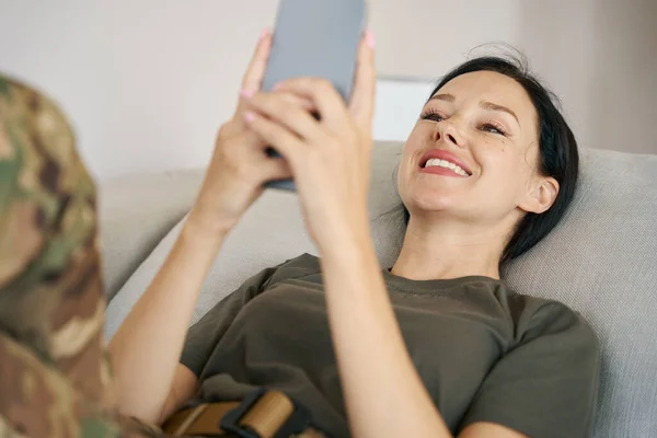 Smiling military woman lies on the couch, holds a mobile phone in her hands and watches content joyfully