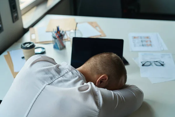 Exhausted consultant in a white shirt fell asleep at the workplace in the office, on his desk there is coffee, a laptop, pencils and glasses