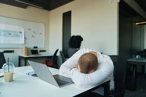 Tired man fell asleep at his desk in the office, in the background a curly woman at her workplace