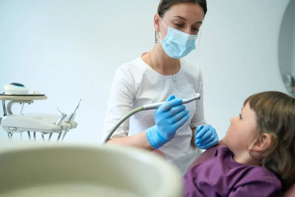 Pediatric dentist in nitrile gloves holding drill handpiece and staring at little patient in chair
