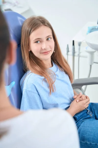 Calm adolescent girl sitting in dental chair and looking at doctor during consultation
