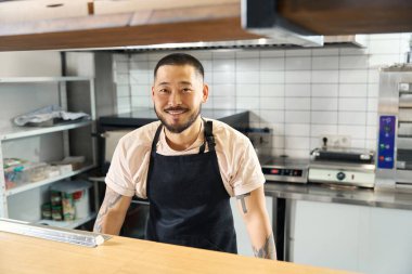 Waist-up photo of excited Asian-looking guy in black apron standing behind cafe counter and looking at camera