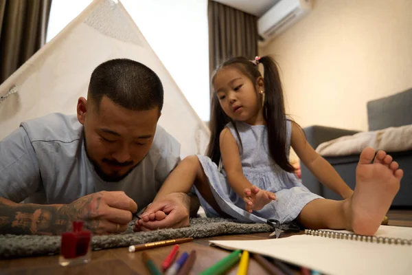 Satisfied asian dad plays with a baby in cozy children room, he paints the toenails of a little serious daughter