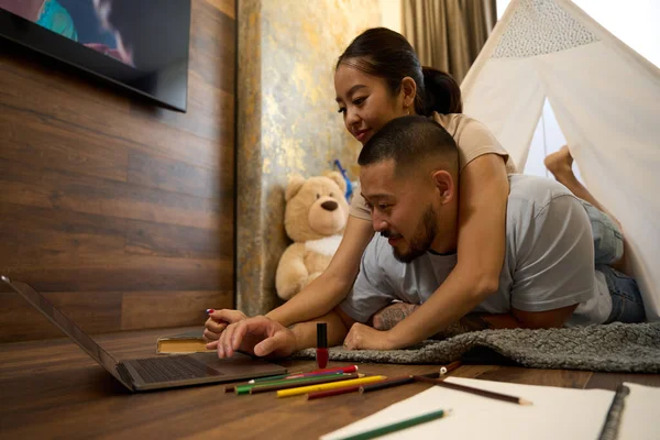 Pretty asian woman lying on back of smiling asian man in kids room in front of laptop on gray mat