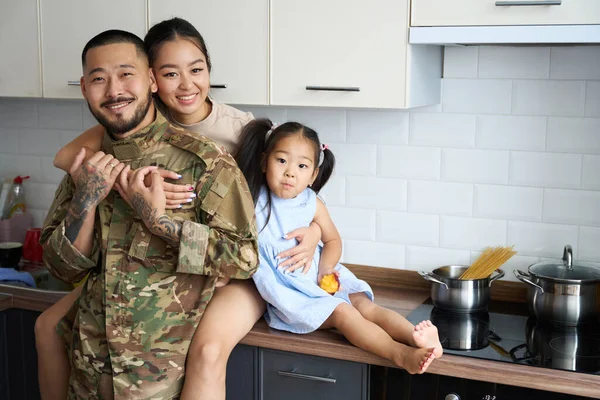 Asian woman hugging military husband in camouflage clothes and pretty little daughter in home kitchen