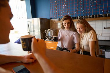 Bir adam karşıda oturmuş elinde bir bardak tutarken neşeli bayan arkadaşıyla barın arkasında duruyor.