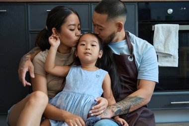 Baba ve anne küçük kızı alnından öpüp kucaklıyor, aile mutfakta yerde samimi.