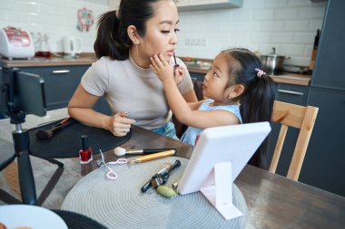 Kameranın önünde sevimli küçük bir kız annesinin dudaklarını özel bir aplikatörle parlatıyor.