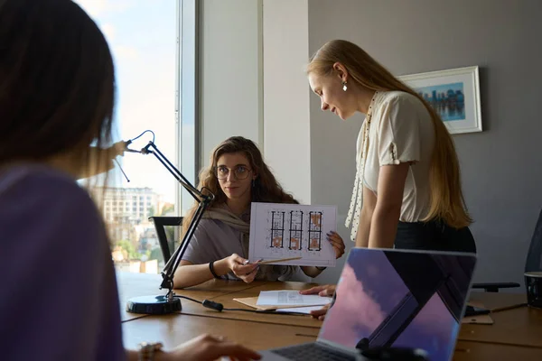 Serious lady sits at the table and stares at a colleague opposite while another one is standing nearby