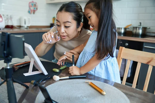Mom asian in the home kitchen in front of the camera teaches her daughter how to curl eyelashes