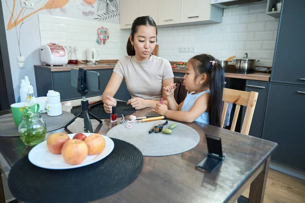 Asian mom blogger in the kitchen on cam teaching beautiful little daughter how to use lip gloss