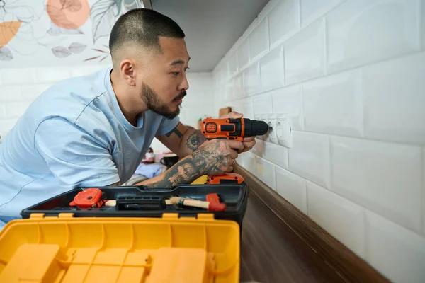 Young asian man is repairing an outlet, he has screwdriver in his hands, a box with a tool stands nearby